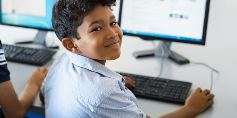 A young boy smiles at the camera while working at his computer in class