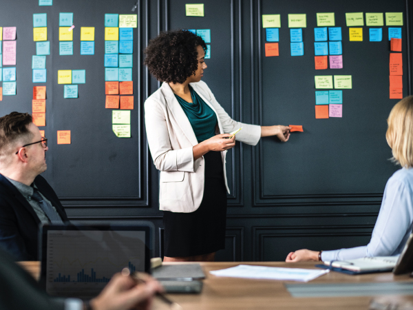 A stock image of someone leading a group discussion using postit notes on a large board / wall