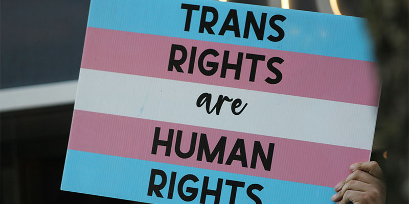 Hand holding a banner showing the Trans Pride flag with the words 'Trans rights are human rights'. Photo by Gabriel Dalton via Unsplash.