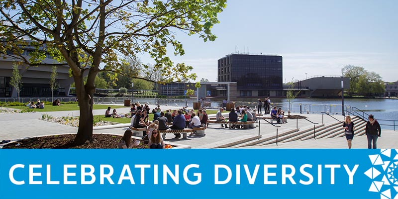 Groups of people gather on Greg's Place with the University lake, Central Hall and the Physics/Electronics building visible in the background. A blue banner with white text reads 'Celebrating Diversity'.