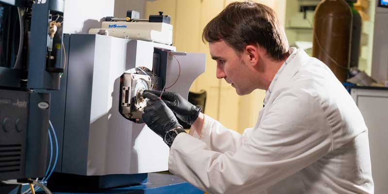 A mian in a white lab coat looks through a microscope