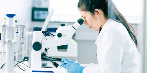 the main image of the Skin centre research web page: a woman looks into a microscope wearing a lab coat and blue nitrile gloves