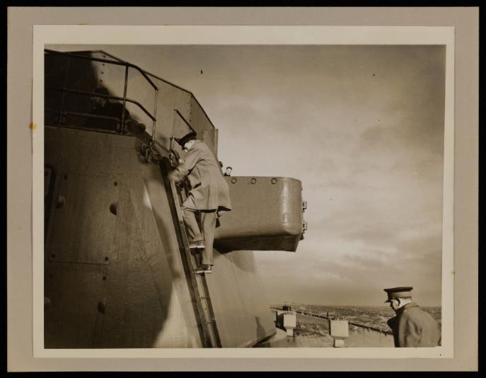 Black and white photograph showing a man in a long coat and black hat climbing down a metal ladder on board a ship.