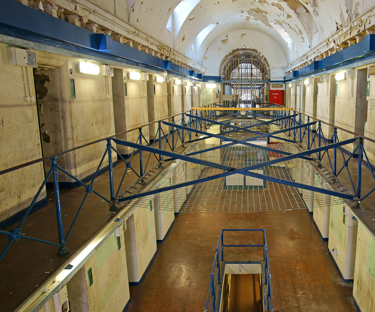 Interior of an abandoned prison. Walkway is central with cell doors to the right and left.