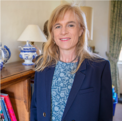 Photograph of Anna Vignoles in blue floral top and dark blue blazer standing in front of bookshelf wiht lamp