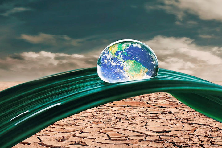 Composite photo image of a parched landscape, with a close up of a blade of grass over it, on which sits a drop of water with an image of the planet earth in it