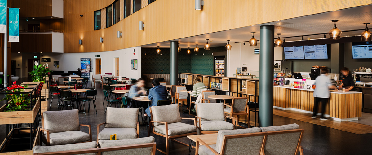 A modern café with cushioned armchairs, wooden tables, and a sleek counter displaying snacks and drinks. Digital menu boards hang above, and pendant lights add warmth. People are seated and ordering, creating a lively atmosphere. The space features greenery and a curved wooden-paneled wall.