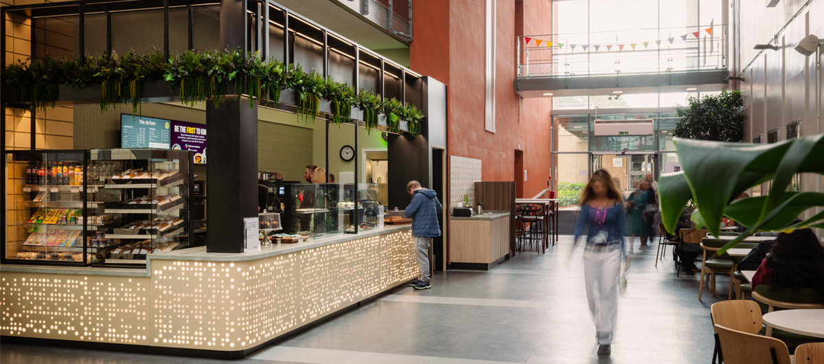A modern indoor café counter with an illuminated, patterned base and hanging greenery stands in a spacious, multi-story atrium as people walk past.