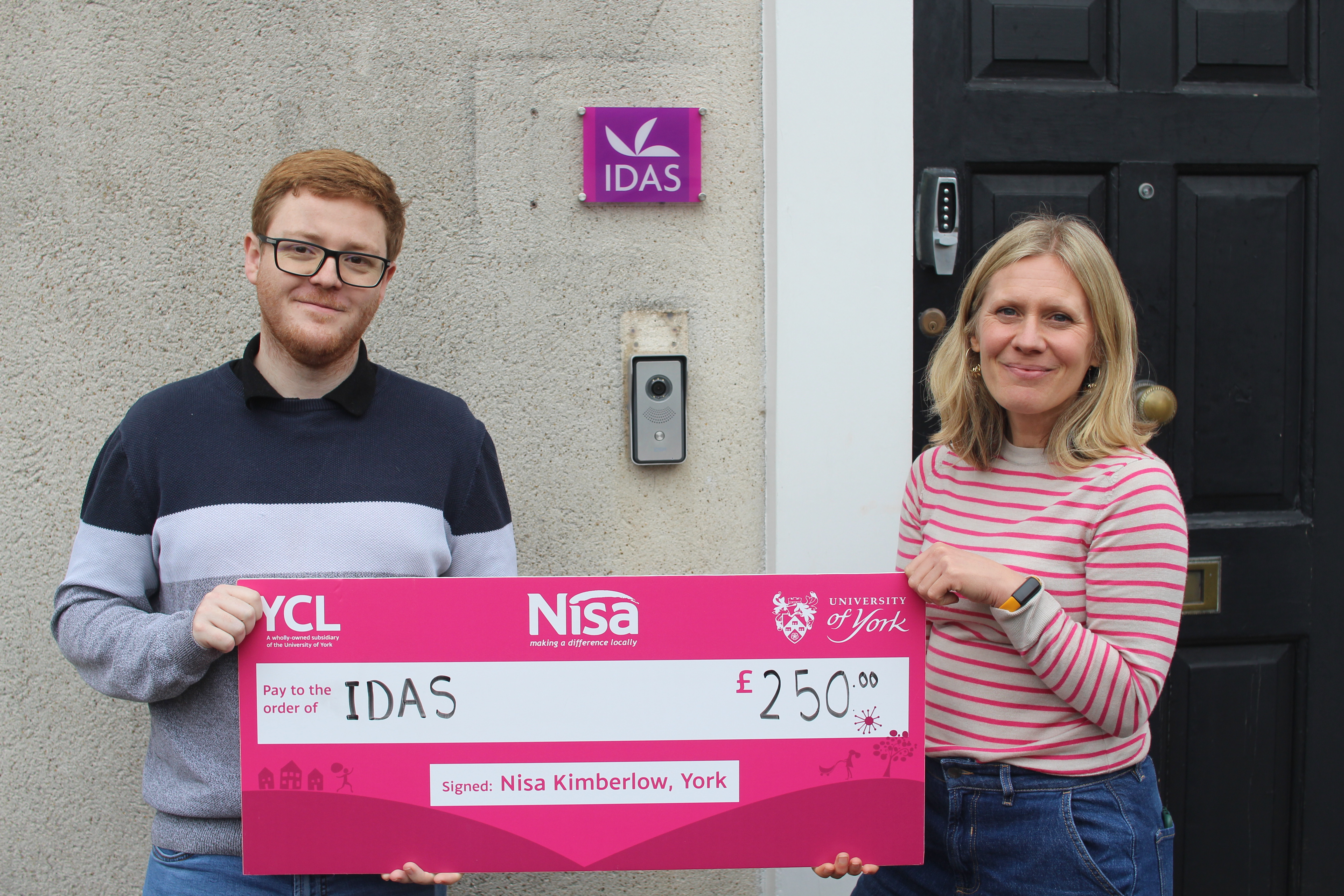 A man from the University of York and a woman from IDAS stand outside an entrance holding a large £250 cheque made out to IDAS, with an IDAS sign on the wall behind them.