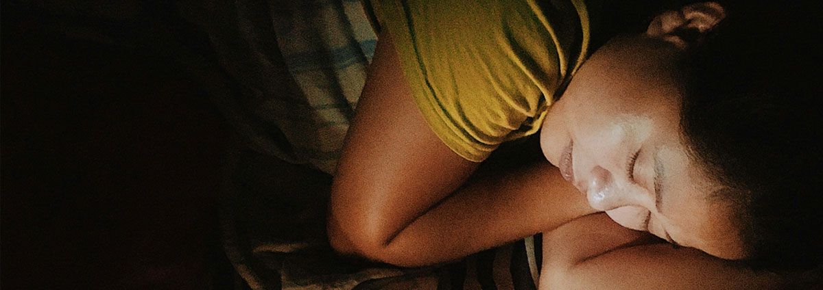 A woman sleeps in a darkened room, her face is visible by the light of a computer screen. Image by Ethan on Unsplash