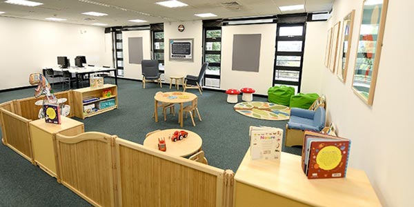 A wide view of the family study room. On the right, there is a children's area with bookshelves, books, toys, colourful beanbags and other children's furniture. On the left, there are several desks and some desktop computers for study.