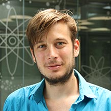Mate Farkas wearing a blue shirt, stood in front of a window, which has a drawing of an atom on.