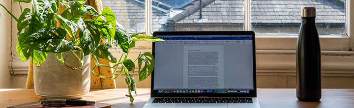 A desk set up for working from home with a laptop, water bottle, notebook and plant.