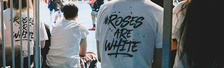 The backs of students watching a basketball game, their T-shirts saying in black on white writing: Roses are White.