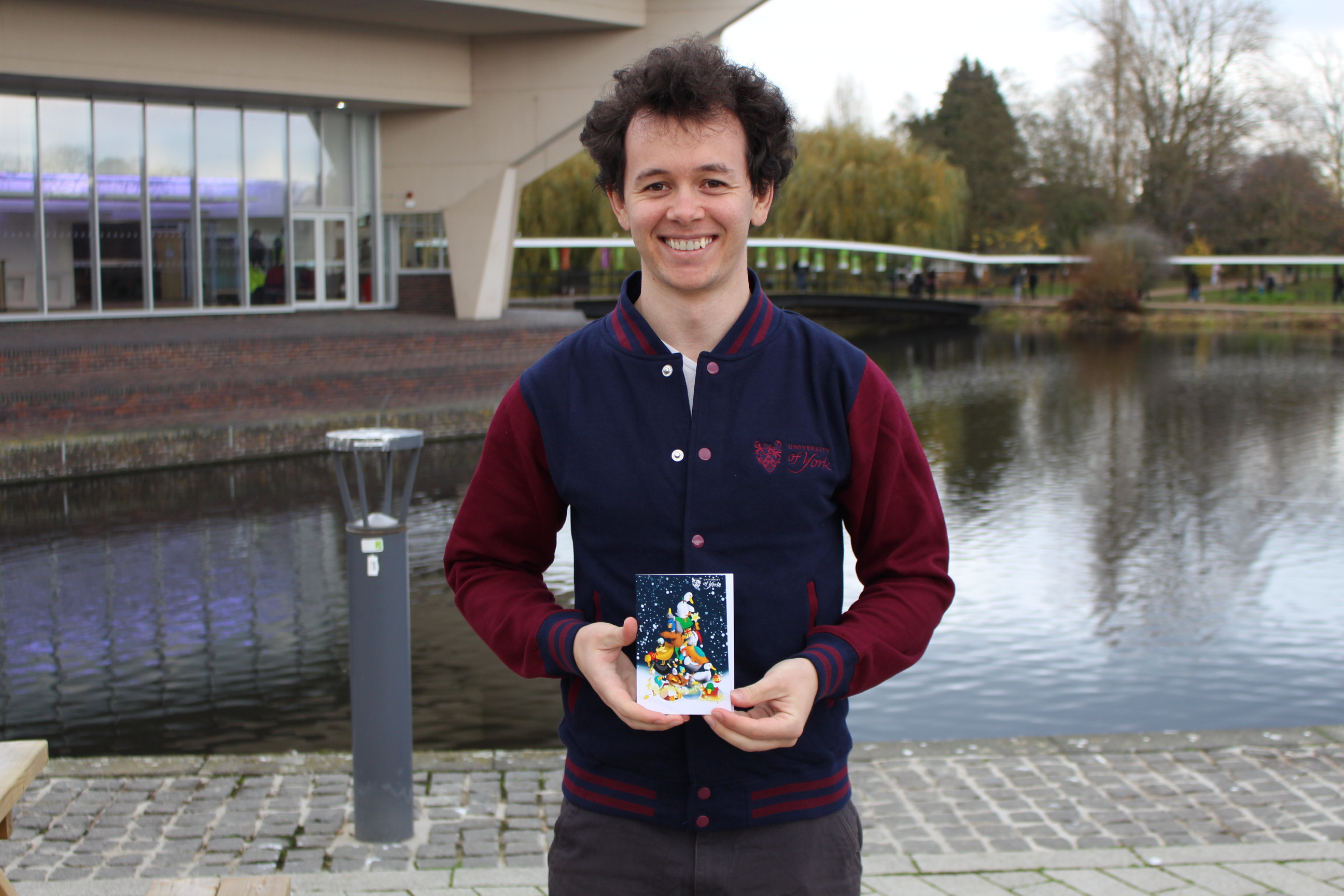A student standing in front of the campus lake, holding his winning Christmas card design. The card features lots of york ducks and geese, wrapped in winter clothes and fairy lights.