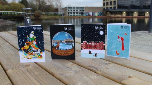 The winning card design and 3 runner-ups are displayed side by side, on a bench in front of the campus lake. Cards feature York waterfowl, central hall in a snowglobe, hes hall in the snow and longboi in a scarf.