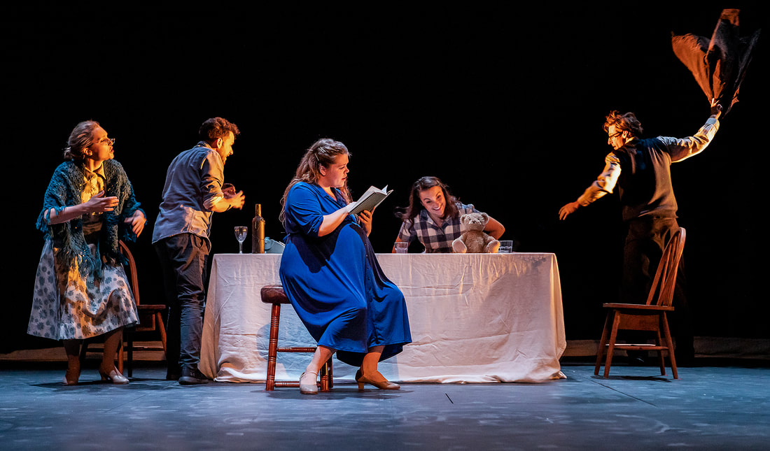 Image: A woman in a blue dress sits in front of a table (center stage) reading a book. To her left at the table, an old woman is stood speaking while a man picks up a glass of wine. To her right a woman sits at the table, smiling at a teddy bear. There is a man stood to the right of the table throwing a jacket across the room. There is warm lighting coming from the right.