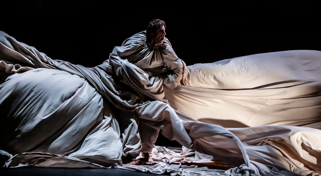 Image: A man is covered in a long strip of cloth that traps him and only shows his head. The cloth is horizontal, going from one side of the stage to the other while he tries to escape it. The floor is also cloth, and the lighting is dim.