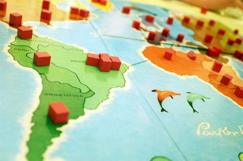 An angled close-up of a vintage Risk board game map showing South America and part of Africa, with small red wooden cube pieces placed on the various territories.