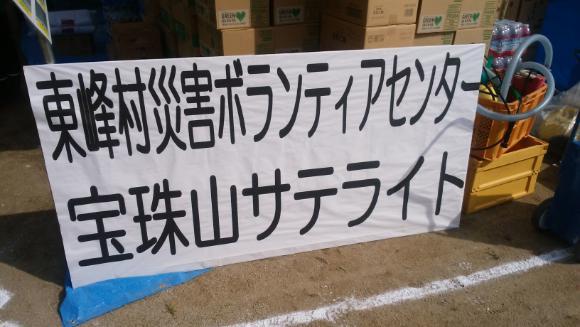 A white banner at an outdoor location, with large black Japanese characters that read: **東峰村災害ボランティアセンター 宝珠山サテライト** (Tōhō-mura Saigai Borantia Sentā Hōshuyama Sateraito), which translates to **Tōhō Village Disaster Volunteer Center Hōshuyama Satellite**.